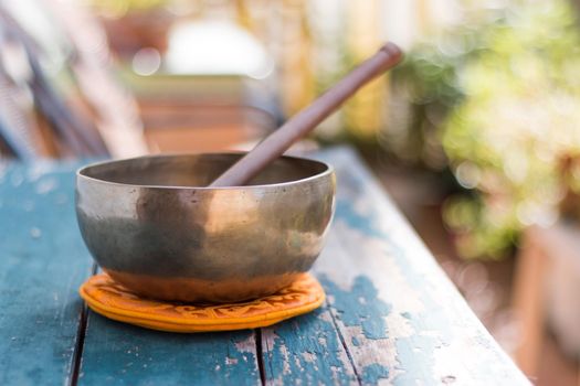 Metal singing bowl on a rustic green, wooden table outdoors. Flowers in the colourful, blurry background