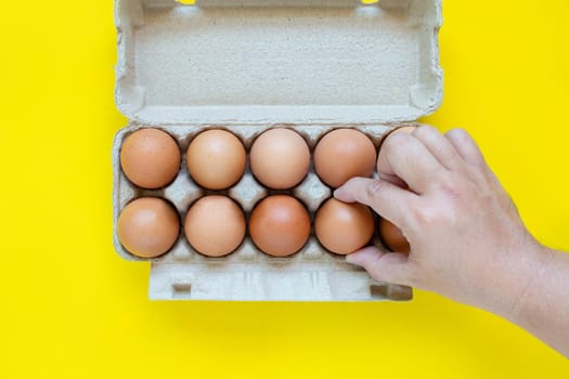 Man's hand is picking up a brown egg in a carton box. On a yellow background.