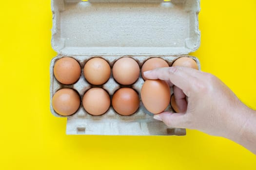 Man's hand is picking up a brown egg in a carton box. On a yellow background.