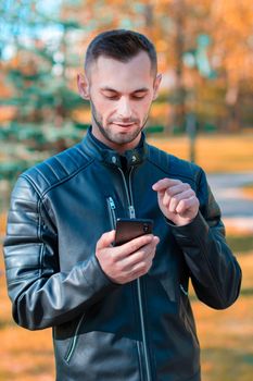 Youthful Guy Using Black Smartphone at the Beautiful Autumn Park. Handsome Young Man with Mobile Phone at Sunny Day - Medium Shot Portrait
