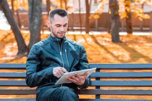 Handsome Young Man Sitting on the Bench and Using Big White Tablet PC at the Beautiful Autumn Park