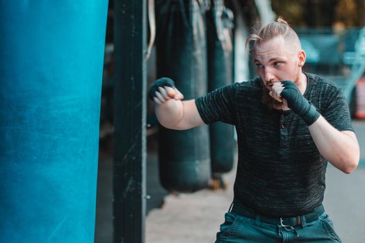 Street Fighter in Black Clothes and Bandages on the Wrist Boxing in Punching Bag Outdoors. Young Man Doing Box Training and Practicing His Punches at the Outside Gym