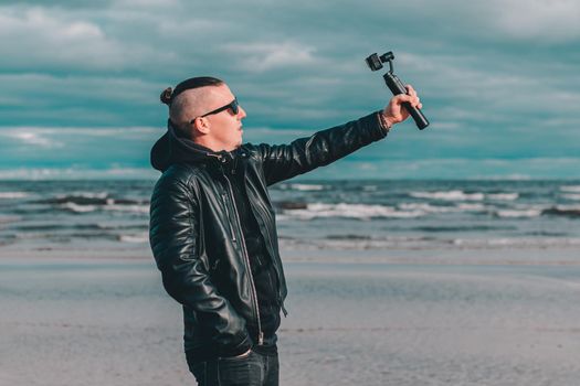 Young Blogger in Sunglasses Making Selfie or Streaming Video at the Beach Using Action Camera with Gimbal Camera Stabilizer. Hipster in Black Clothes Making Photo Against the Sea