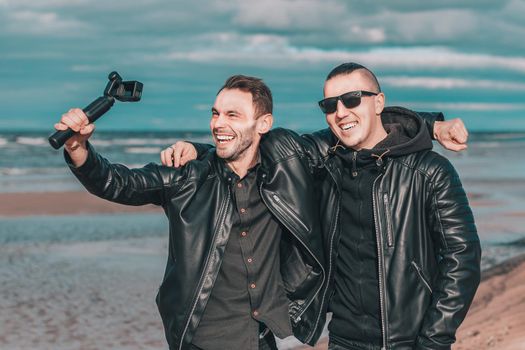 Two Handsome Smiling Friends Making Selfie Using Action Camera with Gimbal Stabilizer at the Beach. Youthful Men in Black Clothes Having Fun by Making Photos