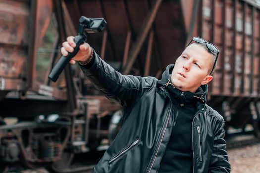 Handsome Guy in Black Clothes and Sunglasses Making Selfie or Streaming Video Using Action Camera with Gimbal Camera Stabilizer at Railway