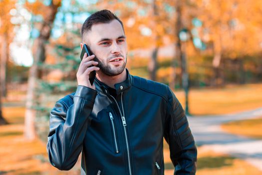 Handsome Young Man Talking on the Phone at the Beautiful Autumn Park. Youthful Guy Using Smartphone for the Call Outdoors at Sunny Day