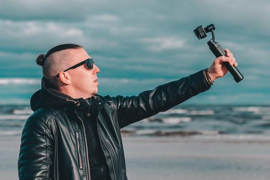 Young Blogger in Sunglasses Making Selfie or Streaming Video at the Beach Using Action Camera with Gimbal Camera Stabilizer. Hipster in Black Clothes Making Photo Against the Sea