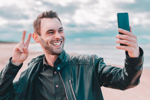 Young Blogger Making Selfie or Streaming Video at the Beach Using Black Smartphone. Handsome Man in Black Clothes Making Photo Against the Sea at Cloudy Day