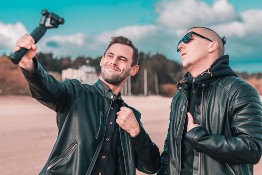 Two Handsome Male Friends Making Selfie Using Action Camera with Gimbal Stabilizer at the Beach. Youthful Men in Black Clothes Having Fun by Making Photos