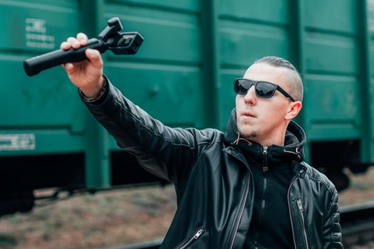 Handsome Guy in Black Clothes and Sunglasses Making Selfie or Streaming Video Using Action Camera with Gimbal Camera Stabilizer at Railway