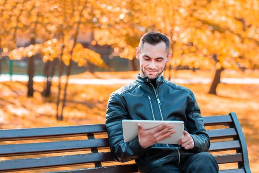 Handsome Young Man Sitting on the Bench and Using Big White Tablet PC at the Beautiful Autumn Park