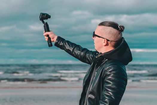 Young Blogger in Sunglasses Making Selfie or Streaming Video at the Beach Using Action Camera with Gimbal Camera Stabilizer. Hipster in Black Clothes Making Photo Against the Sea