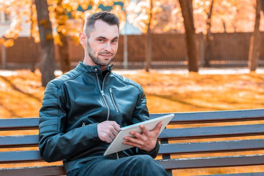 Handsome Young Man Sitting on the Bench and Using Big White Tablet PC at the Beautiful Autumn Park