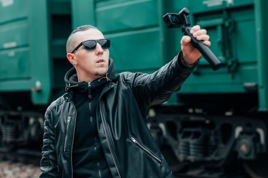 Handsome Guy in Black Clothes and Sunglasses Making Selfie or Streaming Video Using Action Camera with Gimbal Camera Stabilizer at Railway