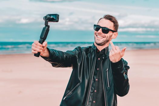 Youthful Blogger in Sunglasses Making Selfie or Streaming Video at the Beach Using Action Camera with Gimbal Camera Stabilizer. Handsome Man in Black Clothes Making Photo Against the Sea