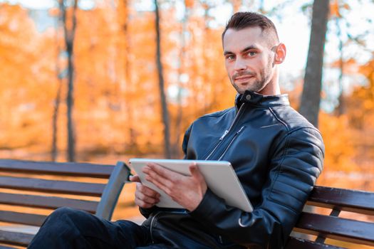 Handsome Young Man Sitting on the Bench and Using Big White Tablet PC at the Beautiful Autumn Park