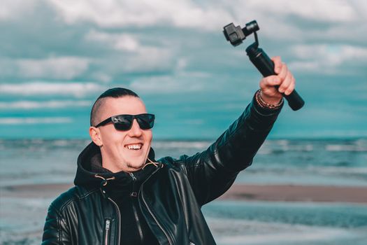 Smiling Young Male Blogger in Sunglasses Making Selfie or Streaming Video at the Beach Using Action Camera with Gimbal Camera Stabilizer. Hipster in Black Clothes Making Photo Against the Sea