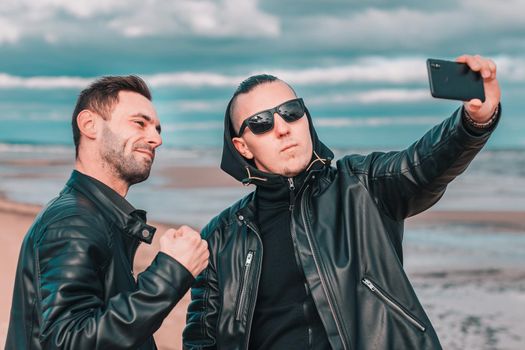 Two Handsome Male Friends Making Selfie Using Black Smartphone at the Beach. Youthful Men in Black Clothes Having Fun by Making Photos Against the Sea