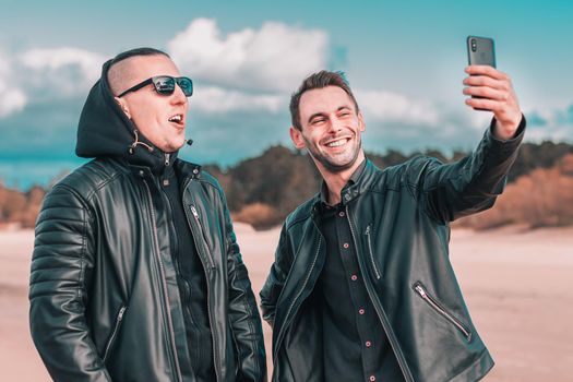 Two Handsome Male Friends Making Selfie Using Black Smartphone at the Beach. Youthful Men in Black Clothes Having Fun by Making Photos Against the Sea