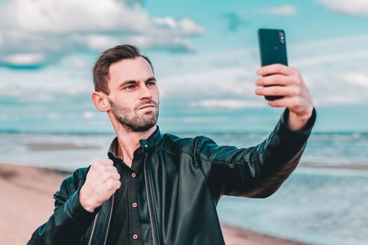 Young Blogger Making Selfie or Streaming Video at the Beach Using Black Smartphone. Handsome Man in Black Clothes Making Photo Against the Sea at Cloudy Day