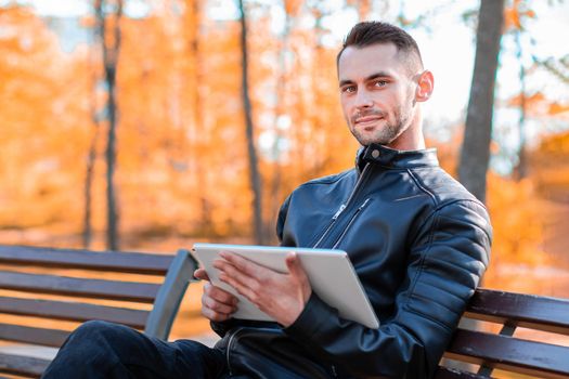 Handsome Young Man Sitting on the Bench and Using Big White Tablet PC at the Beautiful Autumn Park