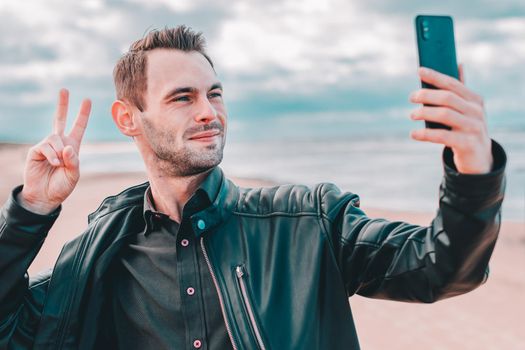 Young Blogger Making Selfie or Streaming Video at the Beach Using Black Smartphone. Handsome Man in Black Clothes Making Photo Against the Sea at Cloudy Day