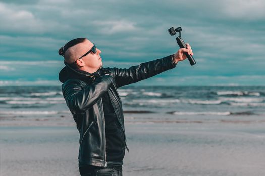 Young Blogger in Sunglasses Making Selfie or Streaming Video at the Beach Using Action Camera with Gimbal Camera Stabilizer. Hipster in Black Clothes Making Photo Against the Sea