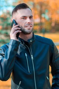 Handsome Young Man Talking on the Phone at the Beautiful Autumn Park. Youthful Guy Using Smartphone for the Call Outdoors at Sunny Day