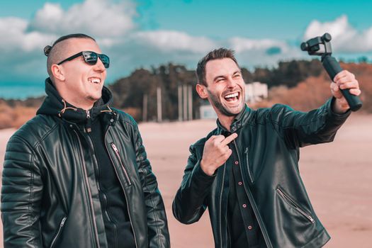 Two Handsome Smiling Friends Making Selfie Using Action Camera with Gimbal Stabilizer at the Beach. Youthful Men in Black Clothes Having Fun by Making Photos