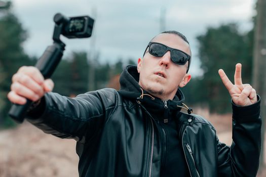 Blogger in Sunglasses Making Selfie or Streaming Video at the Pine Forest Using Action Camera with Gimbal Camera Stabilizer. Handsome Man in Black Clothes Making Photo