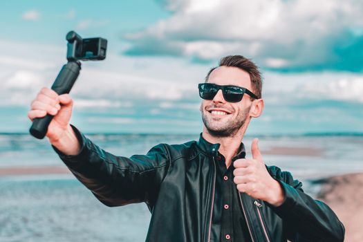 Youthful Blogger in Sunglasses Making Selfie or Streaming Video at the Beach Using Action Camera with Gimbal Camera Stabilizer. Handsome Man in Black Clothes Making Photo Against the Sea