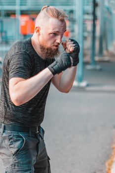 Street Fighter in Black Clothes and Bandages on the Wrist Boxing in Punching Bag Outdoors. Young Man Doing Box Training and Practicing His Punches at the Outside Gym