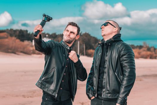 Two Handsome Male Friends Making Selfie Using Action Camera with Gimbal Stabilizer at the Beach. Youthful Men in Black Clothes Having Fun by Making Photos