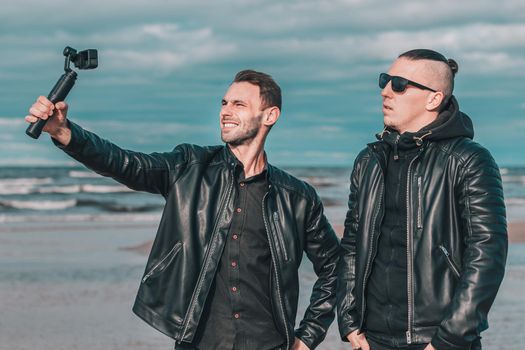Two Handsome Smiling Friends Making Selfie Using Action Camera with Gimbal Stabilizer at the Beach. Youthful Men in Black Clothes Having Fun by Making Photos