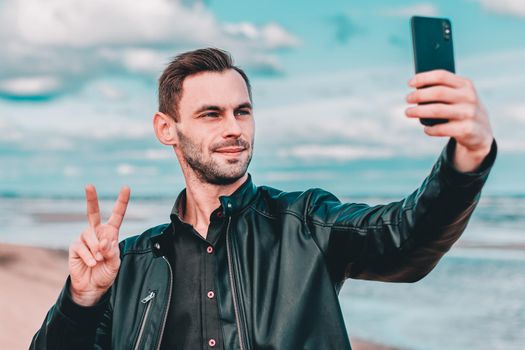 Young Blogger Making Selfie or Streaming Video at the Beach Using Black Smartphone. Handsome Man in Black Clothes Making Photo Against the Sea at Cloudy Day