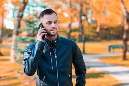 Handsome Young Man Talking on the Phone at the Beautiful Autumn Park. Youthful Guy Using Smartphone for the Call Outdoors at Sunny Day