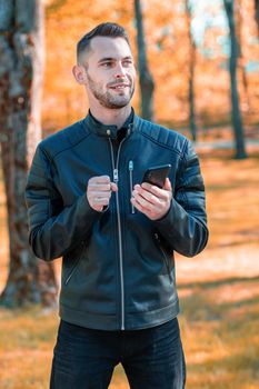 Youthful Satisfied Guy Using Black Smartphone at the Beautiful Autumn Park. Handsome Smiling Young Man with Mobile Phone at Sunny Day - Medium Long Shot