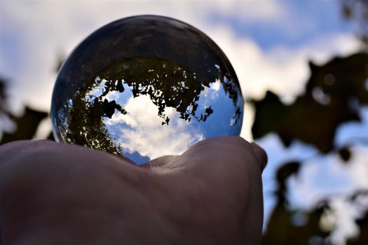 Blue sky with a few clouds and some trees trough a crystal lens ball on a human hand
