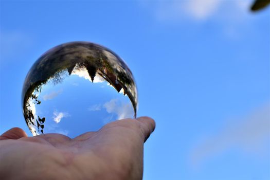 Blue sky with a few clouds and some trees trough a crystal lens ball on a human hand