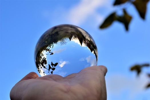 Blue sky with a few clouds and some trees trough a crystal lens ball on a human hand