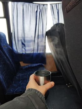 A man holds in his hand a cap from a thermos with steaming hot tea, in the cabin of the bus. Hot drink during the trip.