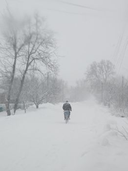 Snow blizzard in the village. A man is riding a bicycle along the street.