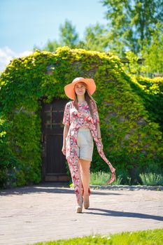 A charming girl in a light summer sundress and a pareo hat is walking in a green park. Enjoys warm sunny summer days.