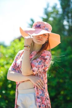 A charming girl in a light summer sundress and a pareo hat is walking in a green park. Enjoys warm sunny summer days.