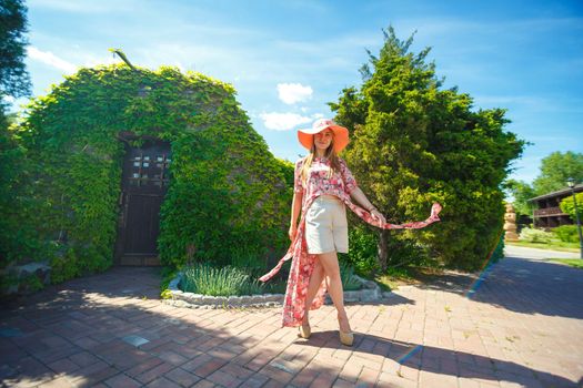 A charming girl in a light summer sundress and a pareo hat is walking in a green park. Enjoys warm sunny summer days.