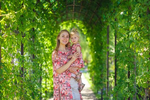 A charming girl in a light summer sundress walks in a green park with her little daughter, holding her in her arms. Enjoys warm sunny summer days.