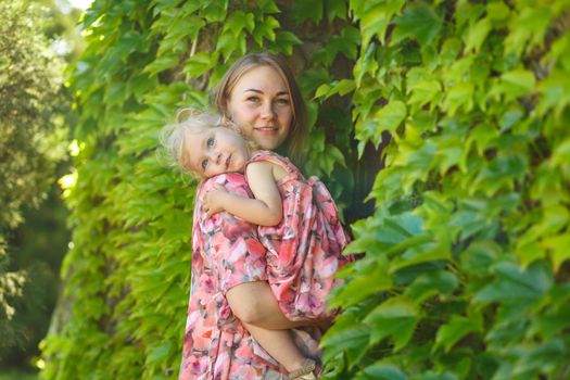 A charming girl in a light summer sundress walks in a green park with her little daughter, holding her in her arms. Enjoys warm sunny summer days.