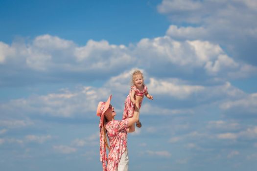 A charming girl in a light summer sundress walks on the sandy beach with her little daughter. Enjoys warm sunny summer days.