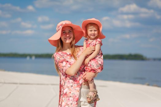 A charming girl in a light summer sundress walks on the sandy beach with her little daughter. Enjoys warm sunny summer days.