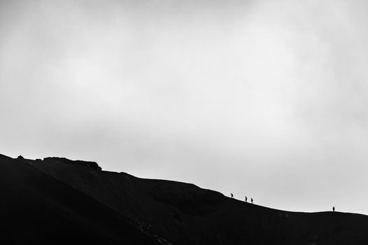 Minimalist image of small silhouette of men climbing on the edge of Etna volcano crater with a cloudy background